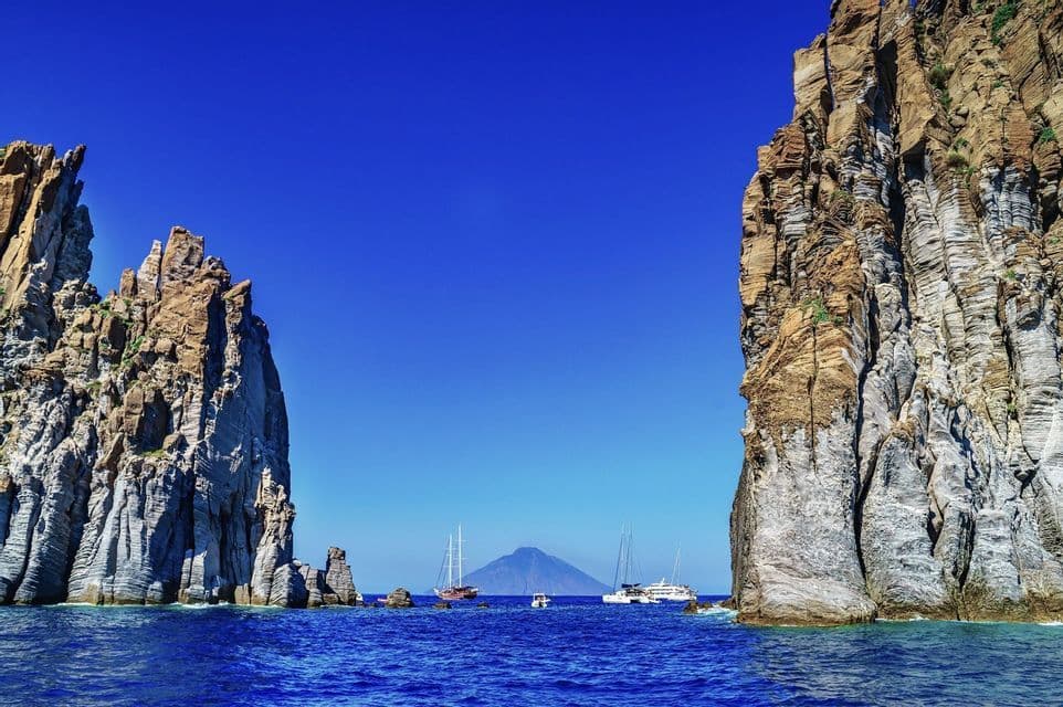 Barche veleggiano su acque azzurre tra due alti faraglioni rocciosi, con un'isola vulcanica distante all'orizzonte.