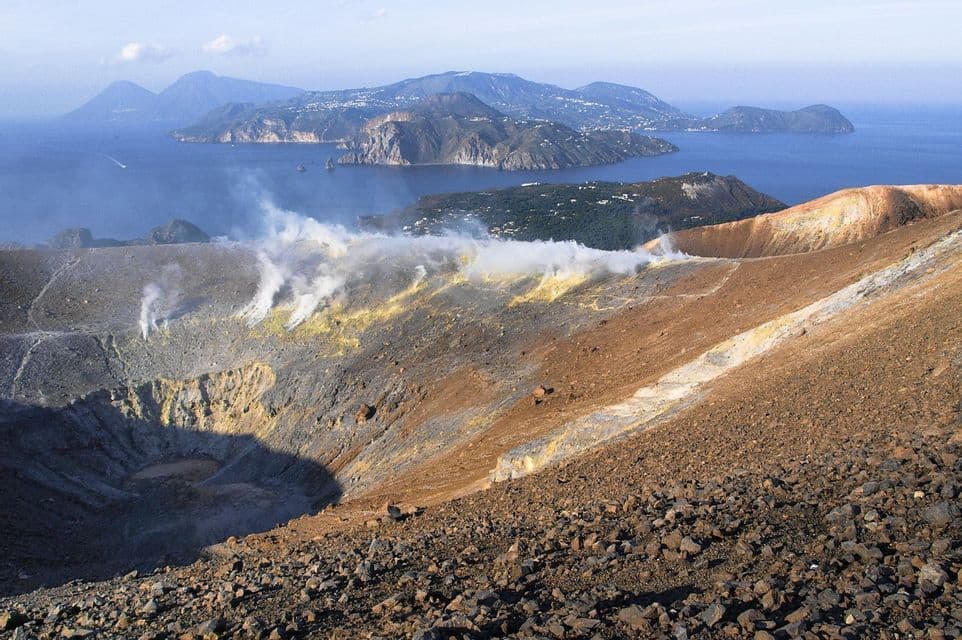 Il vapore sale da un cratere vulcanico che domina isole nel mare, con un pendio roccioso macchiato di zolfo in primo piano.