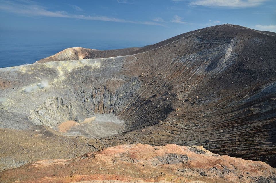Ampia vista di un cratere vulcanico con vapore che si innalza dal suo bordo, affacciato sul mare blu sotto un cielo sereno.