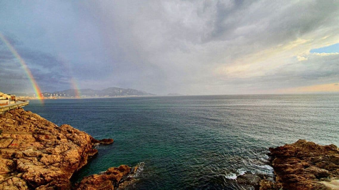 Un double arc-en-ciel s'étend au-dessus de la silhouette d'une ville lointaine, de l'autre côté de la mer, vue depuis un littoral rocheux sous un ciel nuageux.