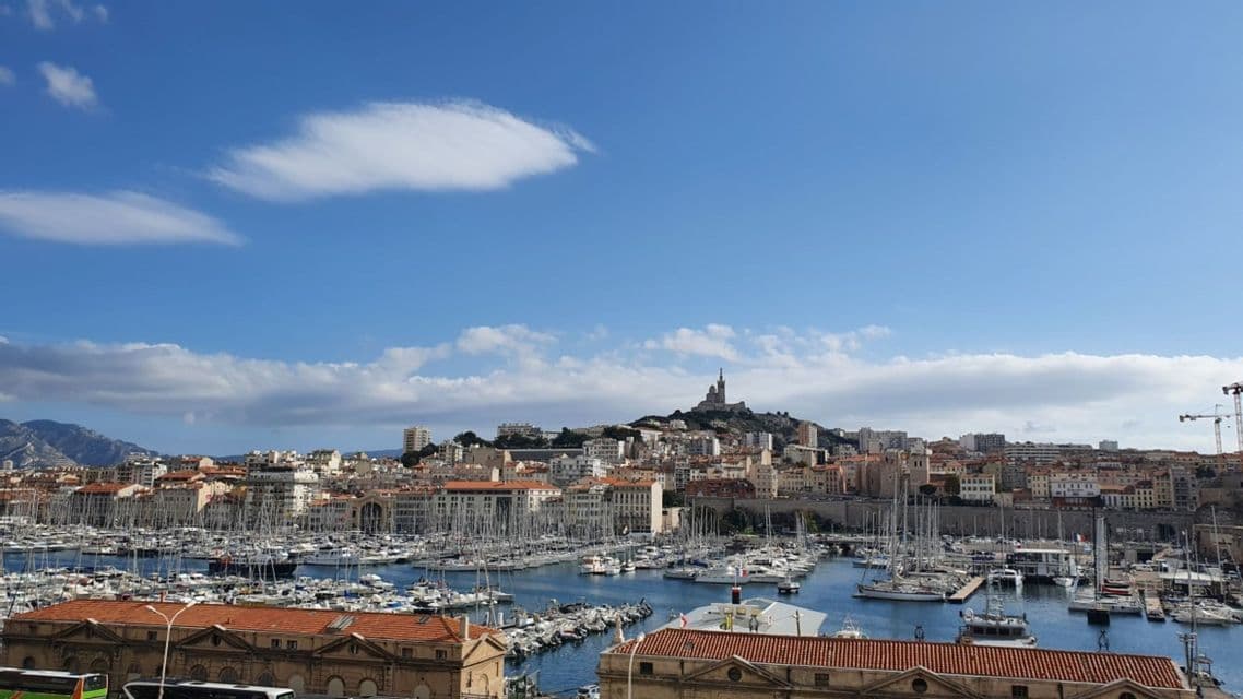 Un grand port rempli de nombreux bateaux amarrés se trouve devant une ville bâtie à flanc de colline, avec une basilique au sommet.
