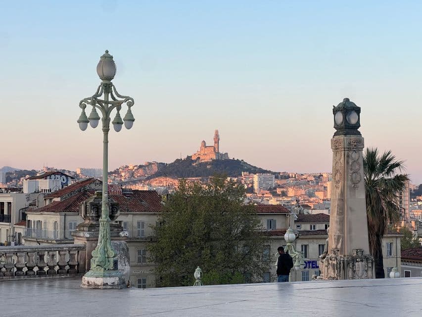 Une personne se tient sur une terrasse avec des lampadaires ornés, admirant un paysage urbain avec une basilique sur une colline sous un ciel de coucher de soleil.