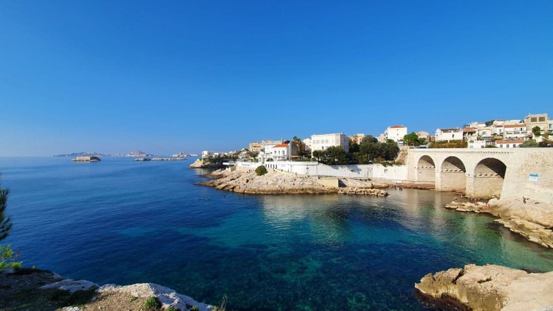 Un pont voûté en pierre et des bâtiments blancs sur une côte rocheuse, à côté d'une mer bleue et limpide, sous un ciel ensoleillé.