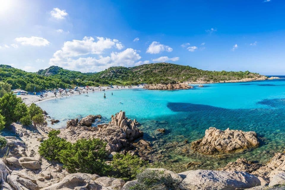 People relaxing and swimming at a secluded beach cove with turquoise water, surrounded by green hills under a sunny sky.