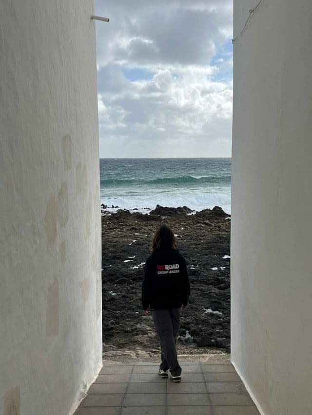 Una persona de espaldas, con una sudadera con capucha de WeRoad Group Leader, camina por un pasaje estrecho hacia una costa rocosa.