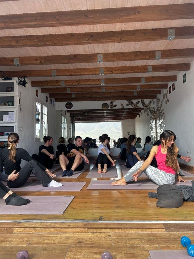 Un gruppo WeRoad in viaggio che partecipa a una lezione di fitness, seduto su tappetini su un pavimento in legno all'interno di uno studio con travi a vista.