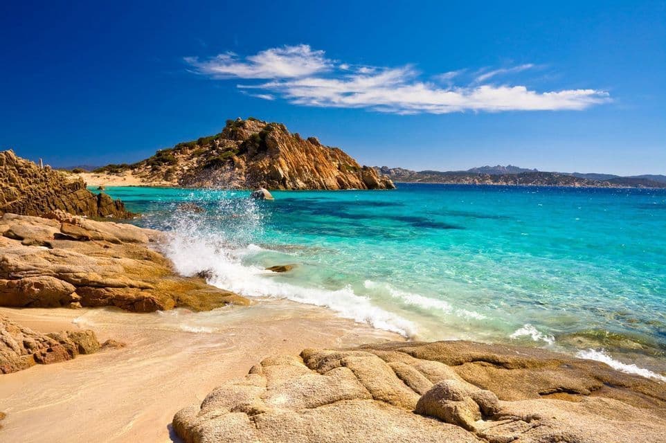 A wave splashes against large rocks on a sandy beach with clear turquoise water under a bright blue sky.