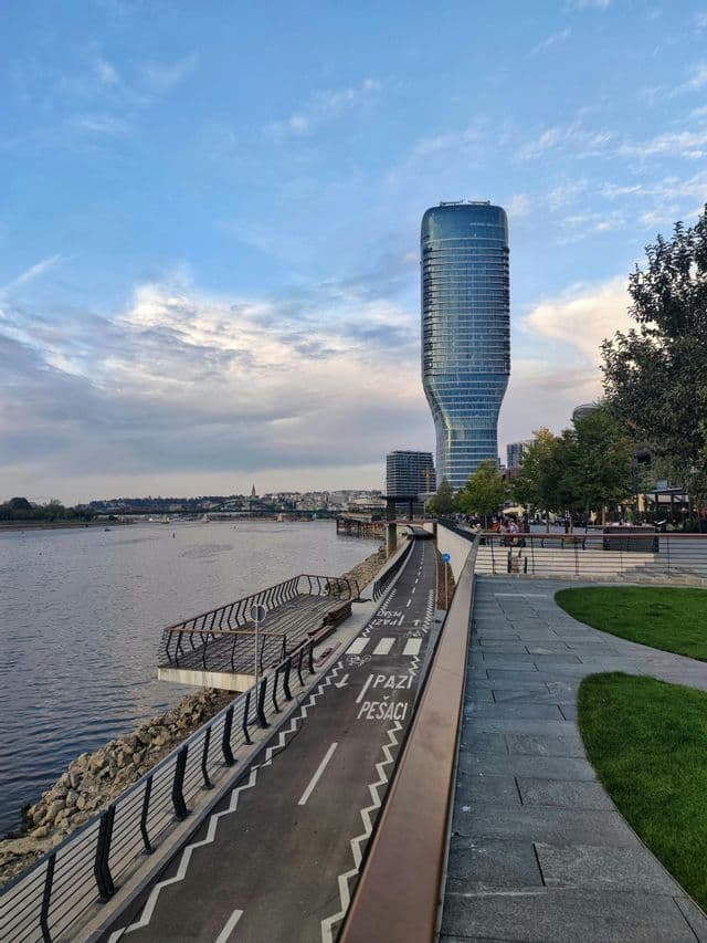 Un moderno rascacielos de cristal curvo domina un paseo fluvial con carril bici. El horizonte de la ciudad es visible al otro lado del agua bajo un cielo nublado.