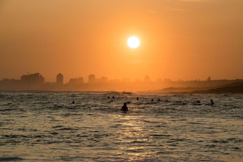 Un gruppo di surfisti in silhouette nell'oceano al tramonto, con il sole e lo skyline nebbioso di una città sullo sfondo.