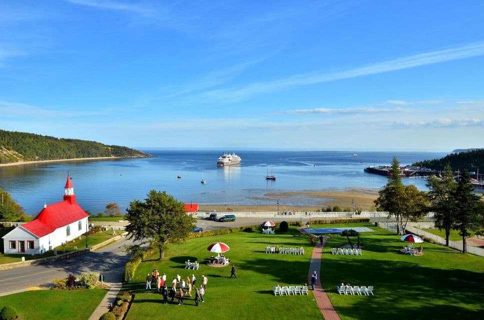 Vue aérienne d'une ville côtière avec une église blanche au toit rouge, une pelouse verte avec des gens et une baie avec un bateau de croisière.