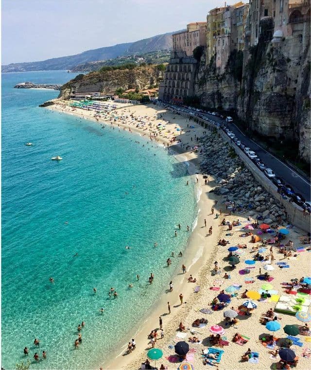 Una vista aerea di una spiaggia sabbiosa affollata con acqua turchese, ombrelloni colorati e una città costruita su una scogliera adiacente.
