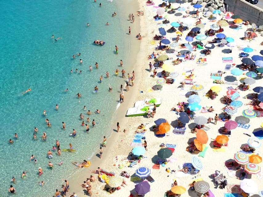 Vista aerea di una spiaggia affollata con molti ombrelloni colorati sulla sabbia bianca e persone che nuotano nell'acqua cristallina turchese.