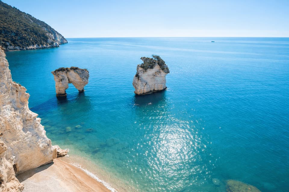 Due faraglioni bianchi, uno che forma un arco naturale, si ergono in acque calme e turchesi accanto a una scogliera e una piccola spiaggia sabbiosa.
