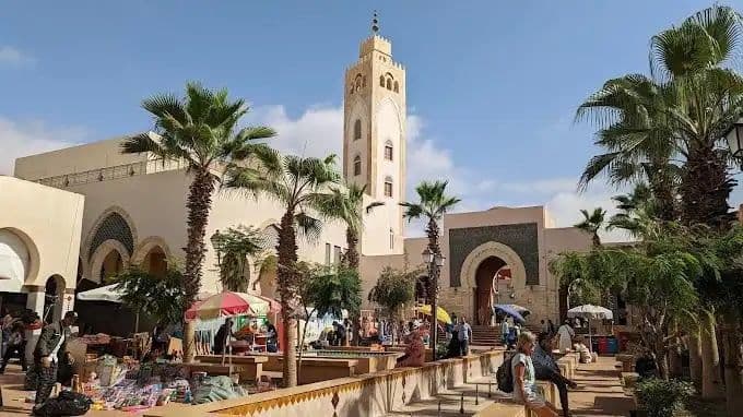 Un marché en plein air grouille de monde dans une cour ensoleillée, avec un grand minaret de mosquée et des palmiers en arrière-plan.