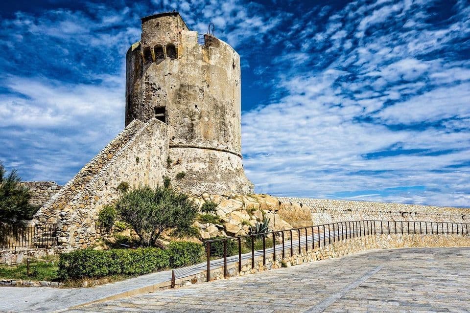 Una torre circolare in pietra, logorata, con una scala esterna, su uno sperone roccioso sotto un cielo azzurro con nuvole bianche.