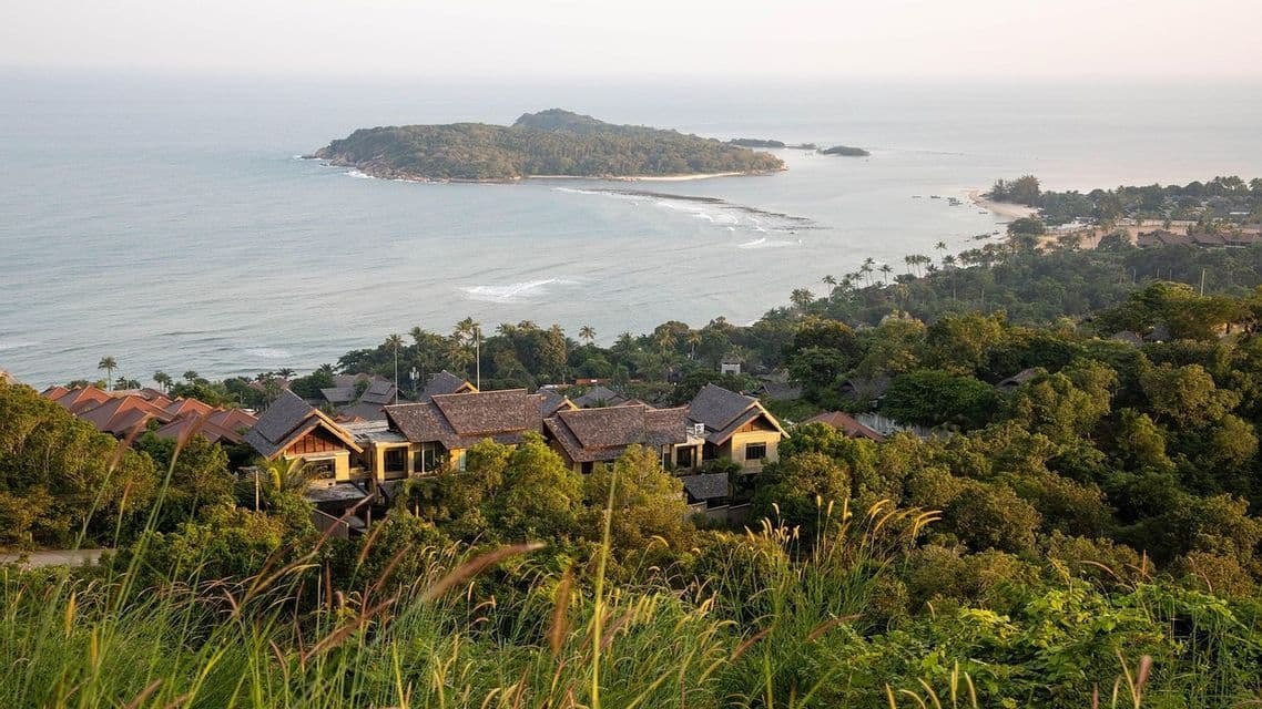 Vue plongeante sur des maisons aux toits de tuiles, nichées dans une forêt tropicale luxuriante, surplombant la mer et une petite île.