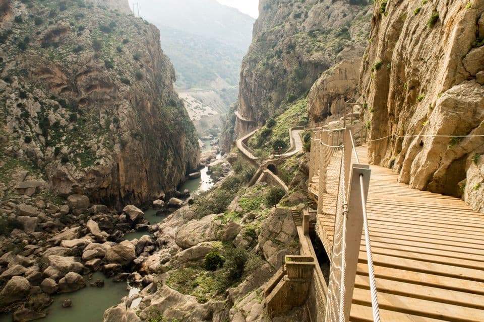 A narrow wooden boardwalk snakes along a sheer rock cliff face high above a river in a deep canyon.