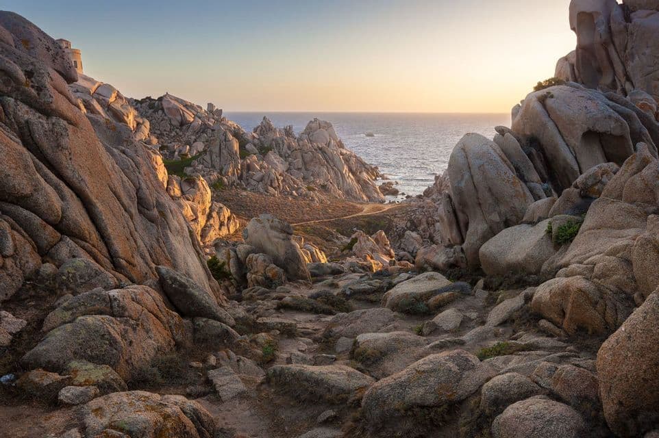 Ein Pfad schlängelt sich durch ein zerklüftetes Tal großer Felsformationen zum Meer, beleuchtet von einem goldenen Sonnenuntergang.