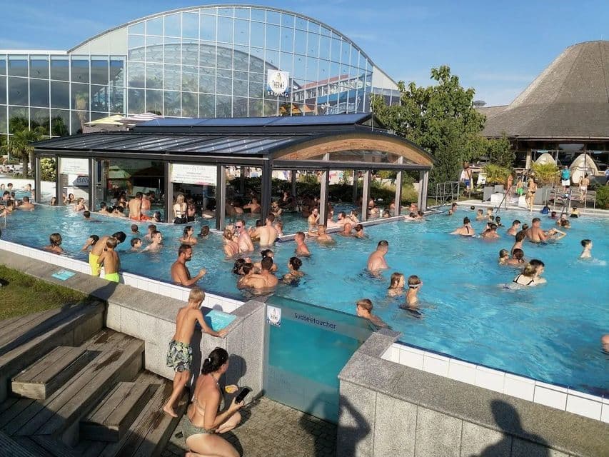 Un folto gruppo di persone nuota e si rilassa in una affollata piscina all'aperto in una giornata di sole, con un moderno edificio in vetro sullo sfondo.