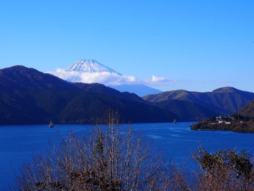 Il Monte Fuji innevato si erge sopra colline boscose e un grande lago blu dove barche navigano sotto un cielo sereno.