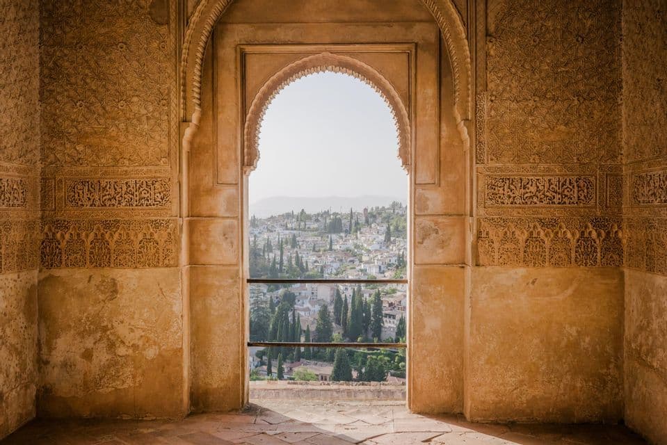 An ornate archway with intricately carved walls frames a sunlit view of a hillside town with white buildings and green trees.