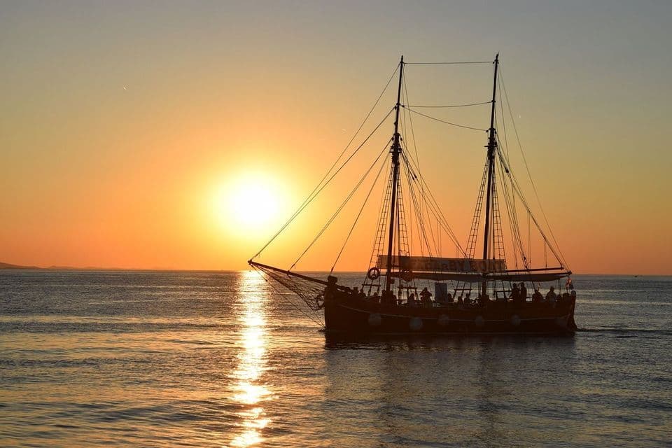 Ein zweimastiges Segelschiff mit einer WeRoad Gruppenreise zeichnet sich vor einem goldenen Sonnenuntergang über dem Meer ab.