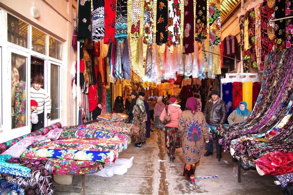 Une allée de marché extérieur animée, bondée de gens qui parcourent des étals remplis de textiles et de tissus colorés.