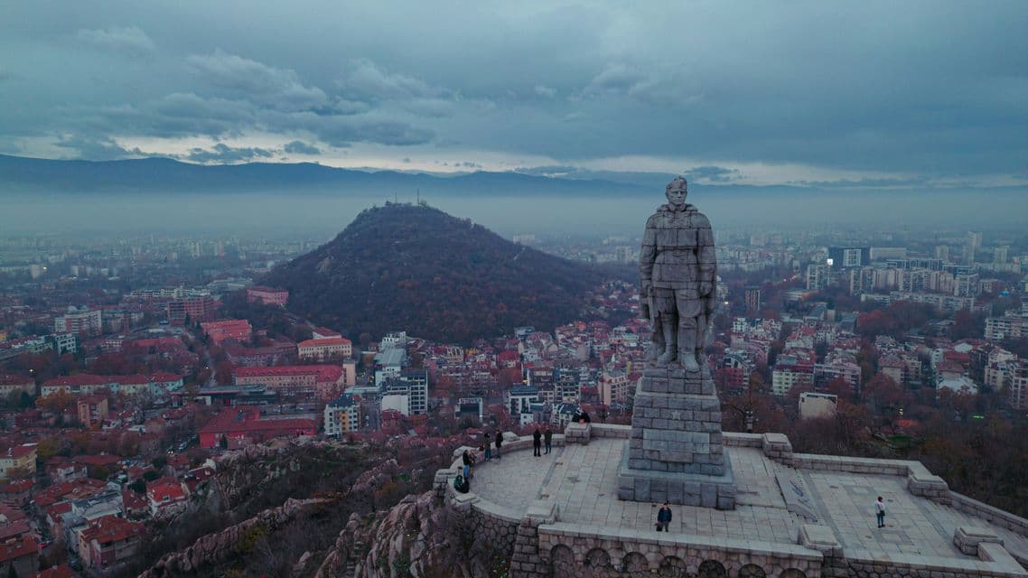 Vue aérienne d'un imposant monument de soldat en pierre, perché sur une colline, dominant une ville tentaculaire sous un ciel nuageux.