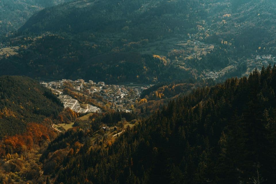 Vue plongeante sur une petite ville nichée dans une vallée montagneuse, entourée de forêts denses de conifères et d'arbres aux couleurs automnales.