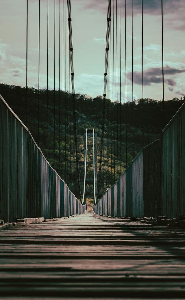 Vue en contre-plongée d'un pont suspendu en bois s'étirant vers une colline verte et boisée sous un ciel nuageux.