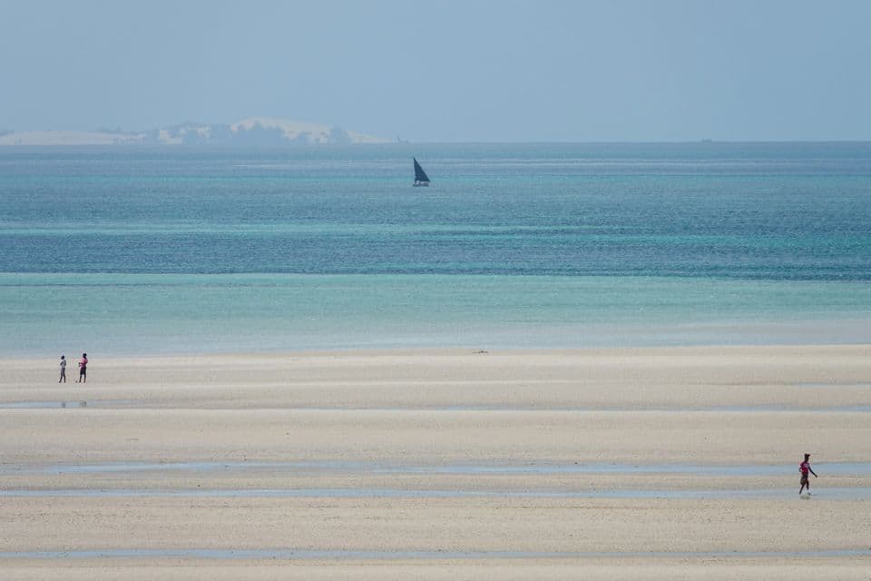 Three people walk on a vast sandy beach at low tide, with a sailboat on the distant blue and turquoise ocean.
