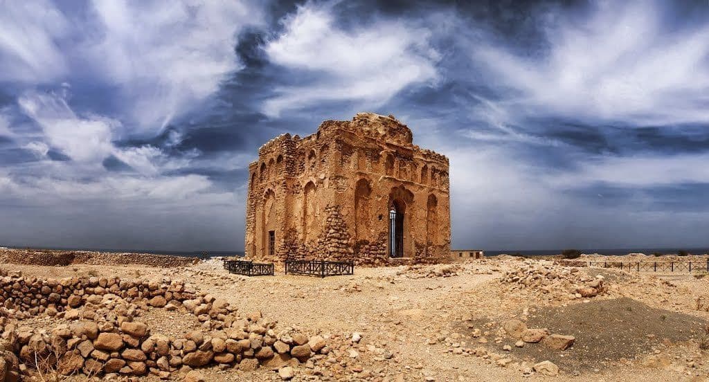 A panoramic view of a large, crumbling stone building with arched details standing in a rocky landscape under a dramatic, cloudy sky.