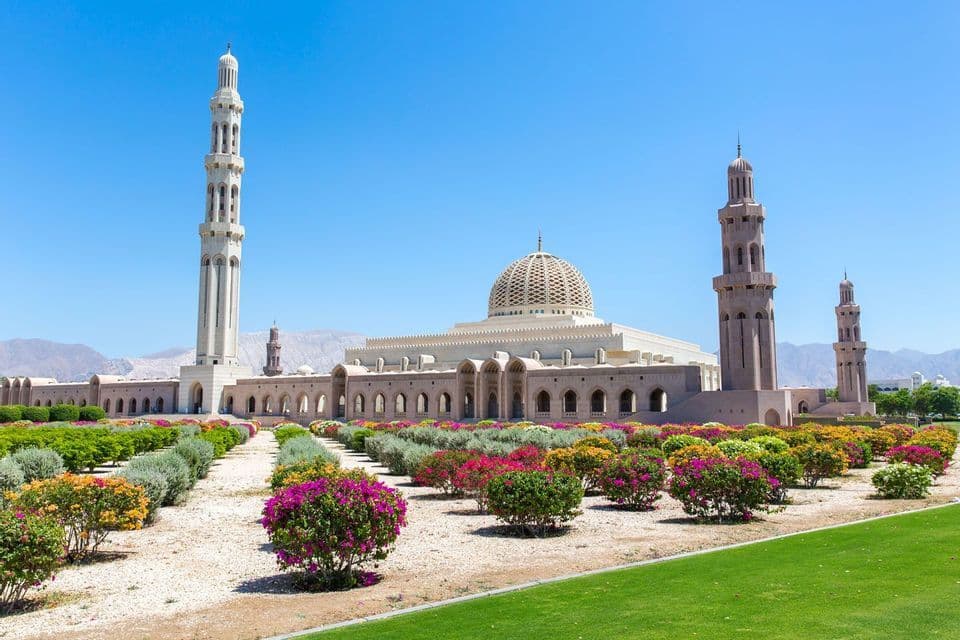 A large mosque with a central dome and several minarets stands behind colorful, manicured gardens under a clear blue sky.