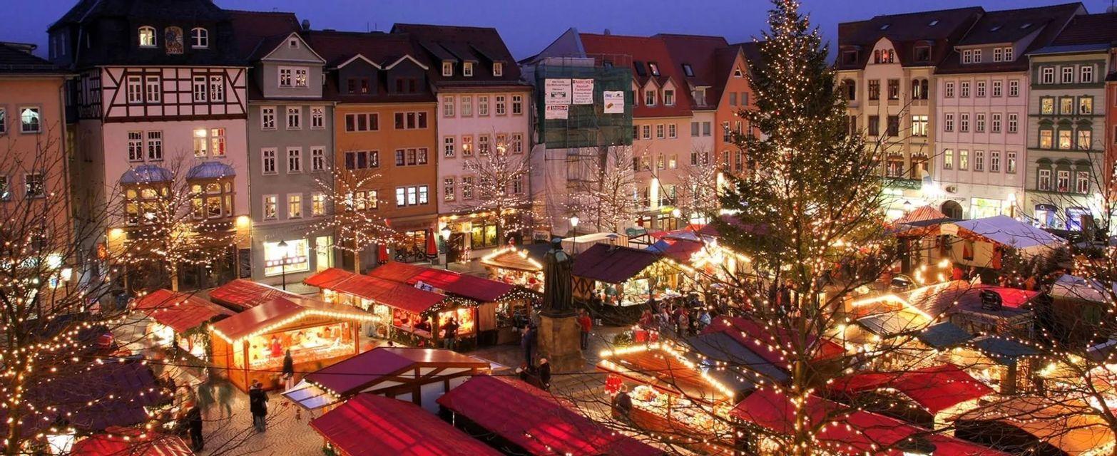 Una vista aerea di un mercatino di Natale in una piazza cittadina al tramonto, con bancarelle e alberi decorati con luci festive.