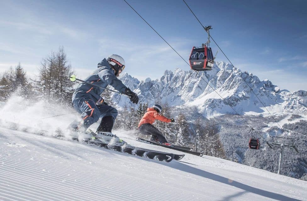 Due persone di un viaggio di gruppo WeRoad sciano su una pista innevata, con una funivia sopra e montagne sullo sfondo.