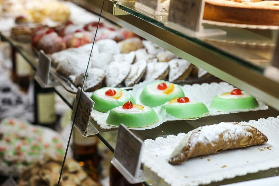 Una varietà di dolci italiani, tra cui un cannolo e cassatine verdi, sono esposti su ripiani di vetro in una vetrina.