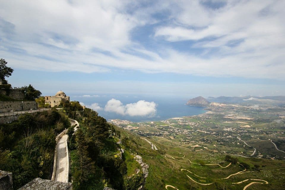 Una vista panoramica da una verde collina, con un edificio in pietra, che domina una città costiera e il mare sotto un cielo parzialmente nuvoloso.