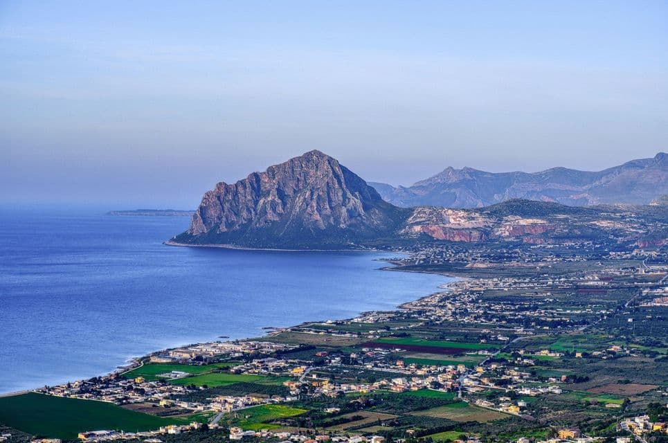 Una vista aerea di una città costiera e campi verdi lungo la riva di un mare blu, con una grande montagna rocciosa sullo sfondo.