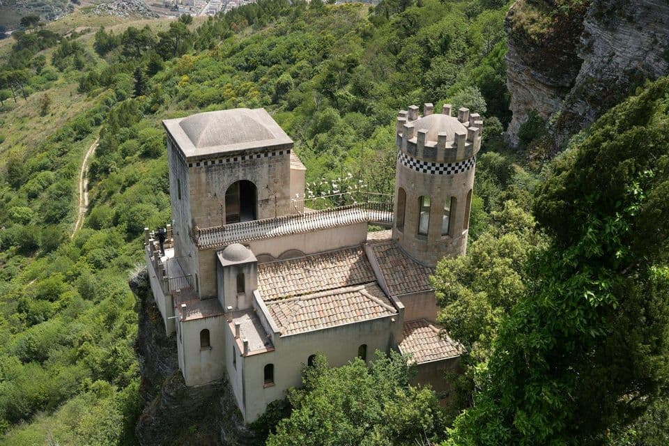 Un antico castello in pietra con una torre quadrata e una torretta rotonda si erge su una scogliera, dominando una rigogliosa foresta verde.