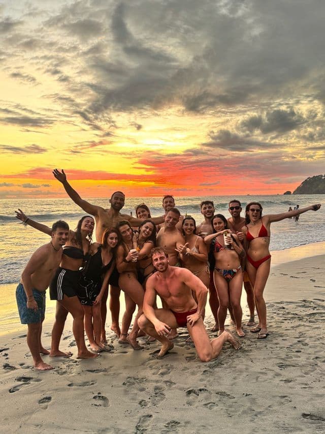 Un gruppo WeRoad in viaggio posa insieme per una foto su una spiaggia sabbiosa durante un tramonto colorato sull'oceano.