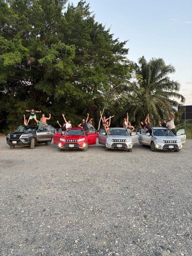 Un gruppo WeRoad in viaggio che esulta mentre posa sopra e intorno a quattro auto parcheggiate in un piazzale ghiaioso con alberi rigogliosi sullo sfondo.