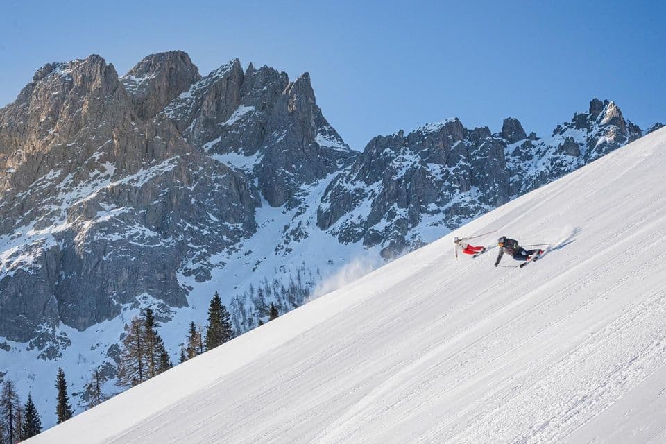 Due sciatori scendono un ripido pendio innevato con montagne rocciose e frastagliate che si ergono sullo sfondo sotto un cielo azzurro e limpido.