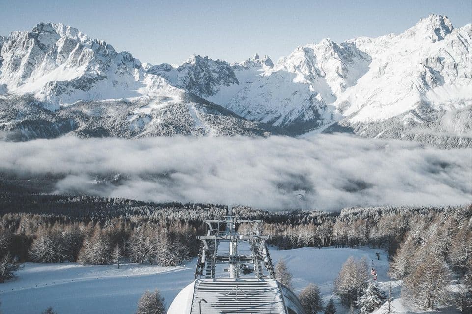 Vista da una stazione di risalita su una valle innevata, con fitti boschi, nuvole basse e imponenti vette montane.