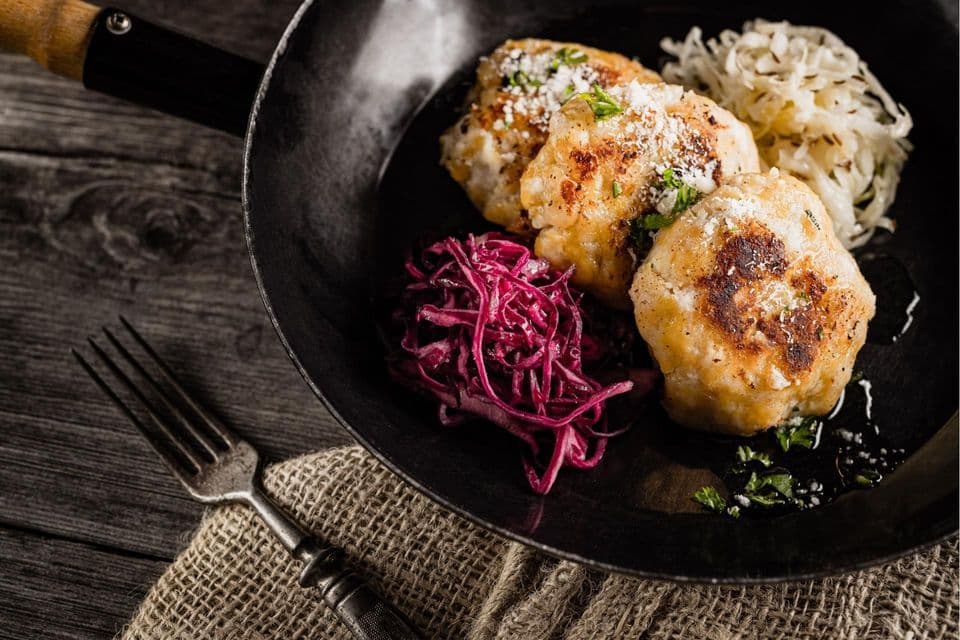 Three pan-fried dumplings in a black skillet with red cabbage and sauerkraut on a rustic wooden surface.
