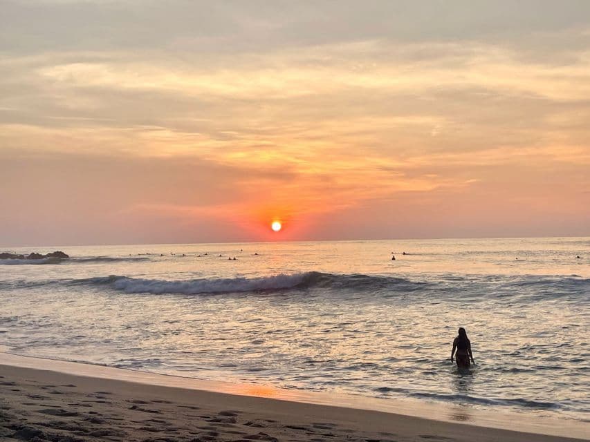 Une personne patauge dans l'océan sur une plage tandis que des surfeurs sont assis sur leurs planches au loin sous un coucher de soleil coloré.