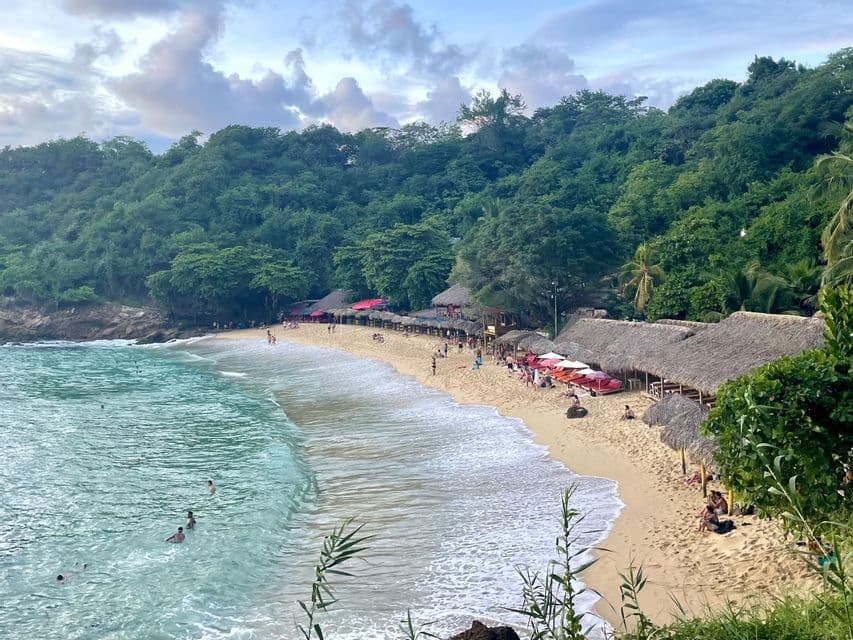 Vue aérienne d'une crique de plage de sable avec des gens qui nagent, des cabanes aux toits de chaume et une forêt verte dense sur une colline.