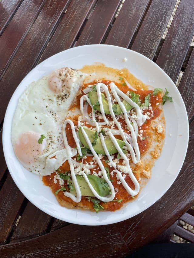 Vue plongeante d'une assiette de chilaquiles à la sauce rouge, garnis d'avocat, de fromage et de crème, servis avec deux œufs au plat.