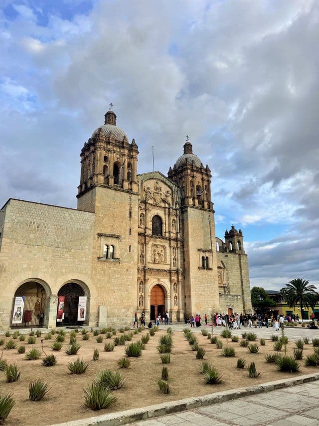 Une église en pierre ornée, dotée de deux clochers, vue depuis une place avec de petites agaves, sous un ciel nuageux.