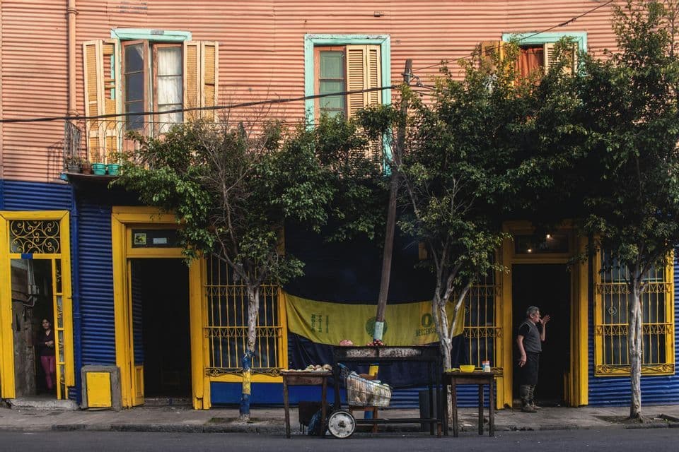 Un edificio colorato con rivestimento in lamiera ondulata rosa e blu, portoni gialli e alberi lungo il marciapiede.