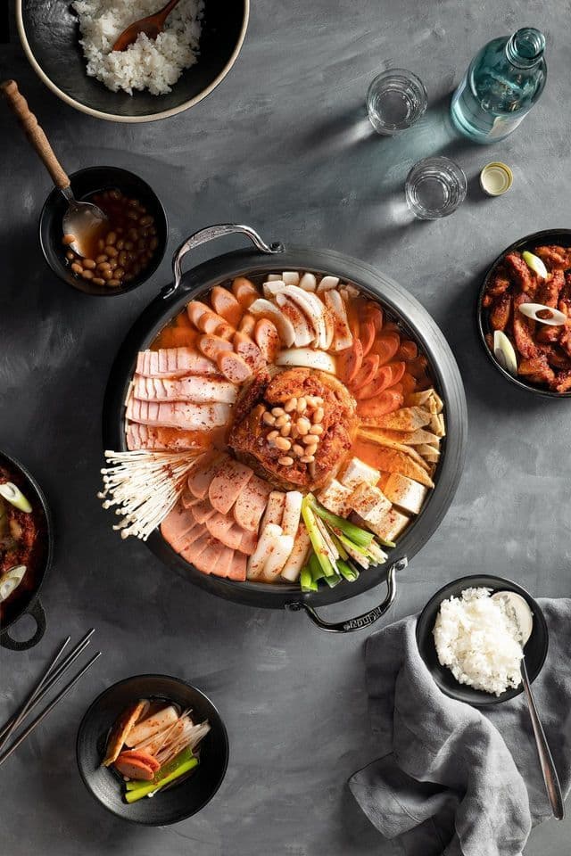 Una vista dall'alto di un hot pot coreano Budae Jjigae, con salsicce, spam, tofu e funghi, circondato da riso e contorni.